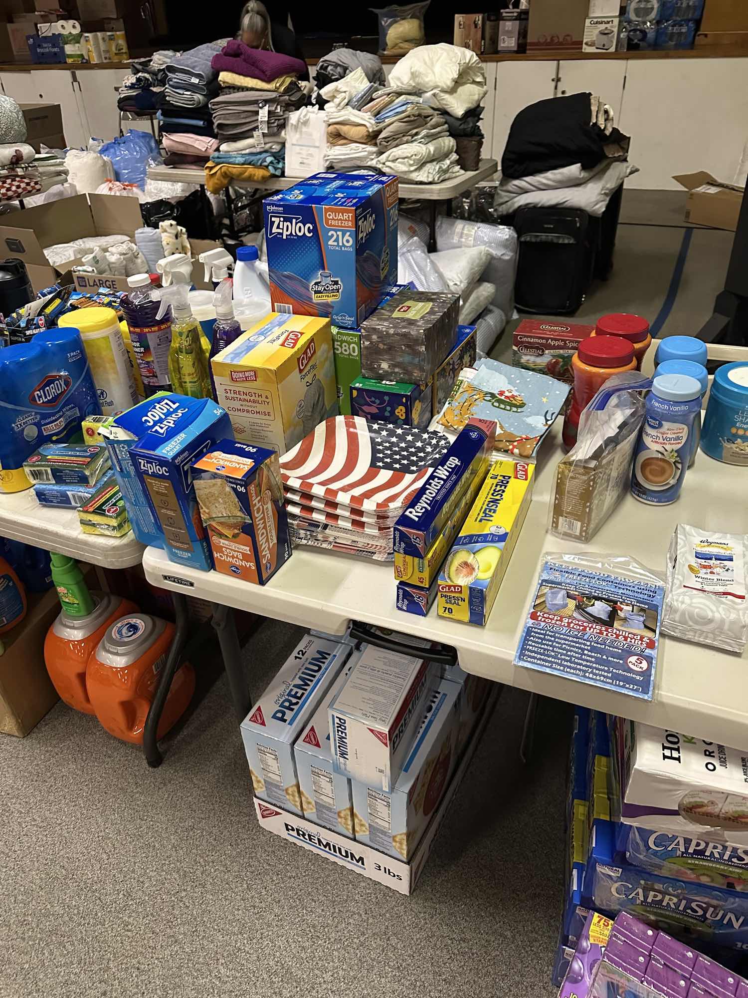 A folding table holding disinfecting wipes, spray cleaners, sponges, laundry detergent jugs and boxes of freezer and sandwich bags, with stacks of folded blankets and towels on tables behind it