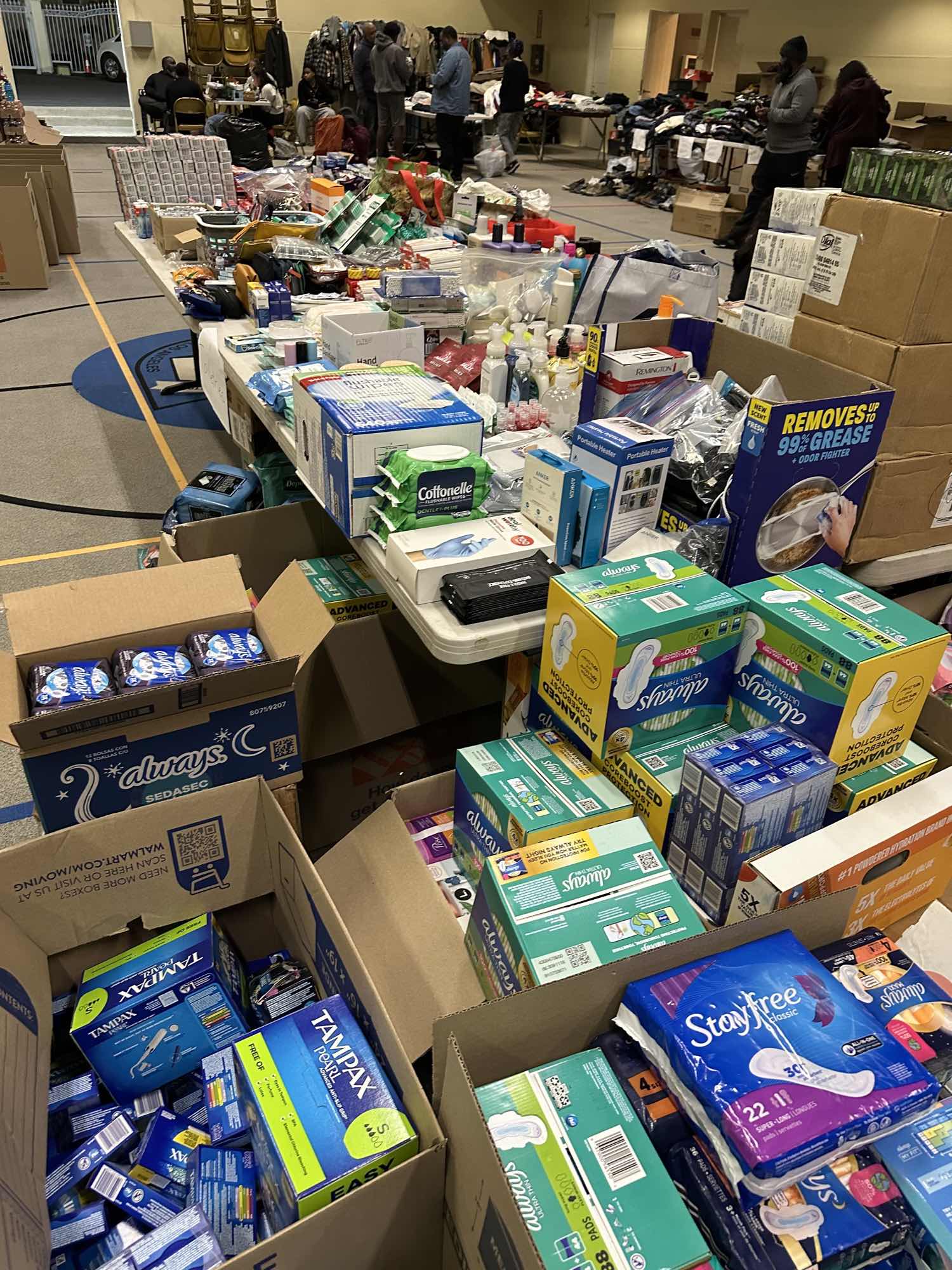 Open cartons of menstrual pads and tampons in the foreground, with a long table behind them holding flushable wipes, hand sanitizer, disposable gloves, toiletries and boxed portable heaters, and people browsing clothing tables in the background