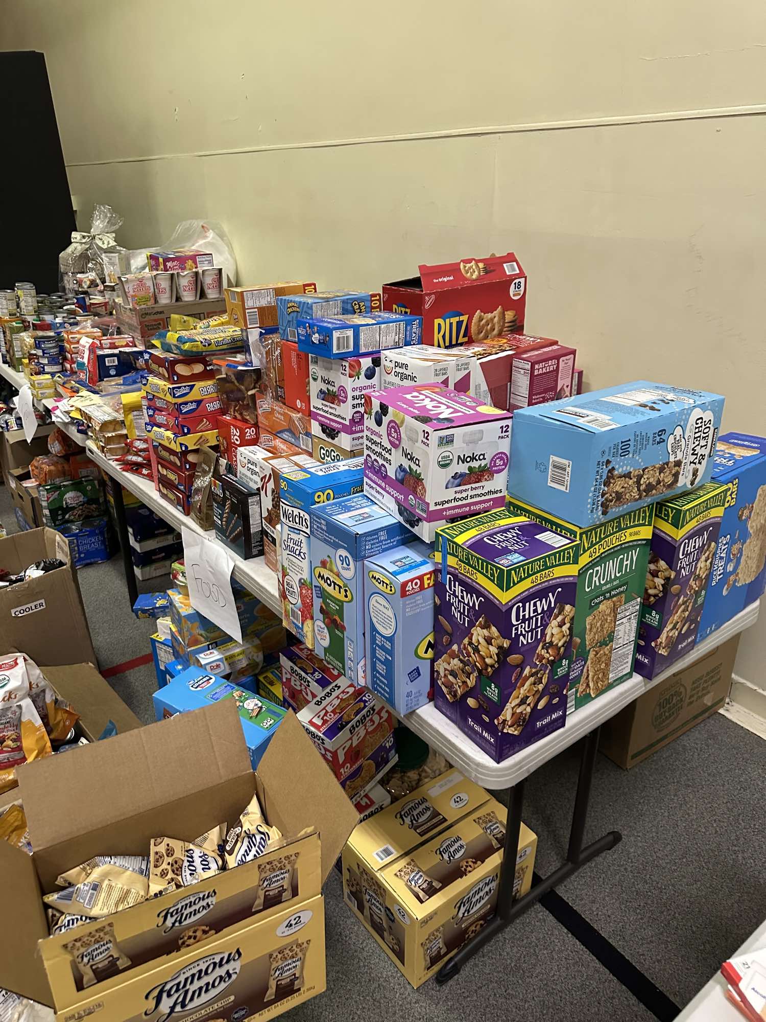 Long tables of shelf-stable food, including canned goods, crackers, granola bars, fruit snacks and instant noodles, with a hand-lettered paper sign reading food and open cartons of packaged cookies on the floor