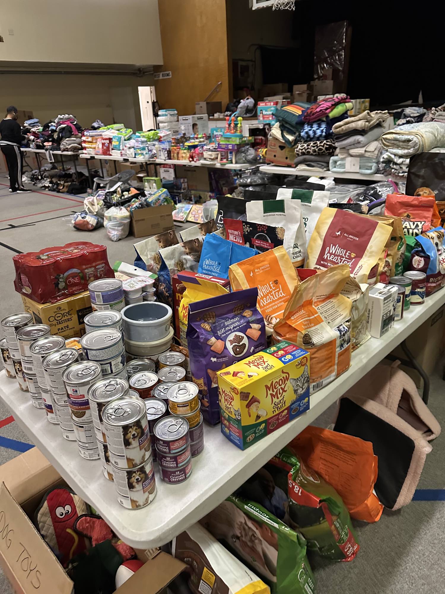 A table of tinned dog and cat food stacked in rows beside bags of dry pet food, pet bowls and a box hand-labeled dog toys, with tables of blankets and clothing in the background