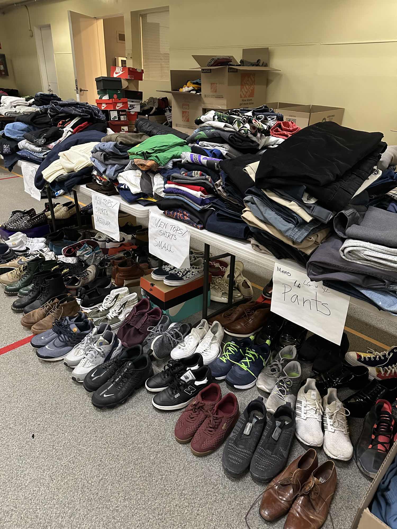Folding tables stacked with folded shirts and trousers behind hand-lettered paper signs reading men's tops shirts medium, men's tops shirts small and men's pants, with dozens of pairs of shoes lined up in rows on the floor