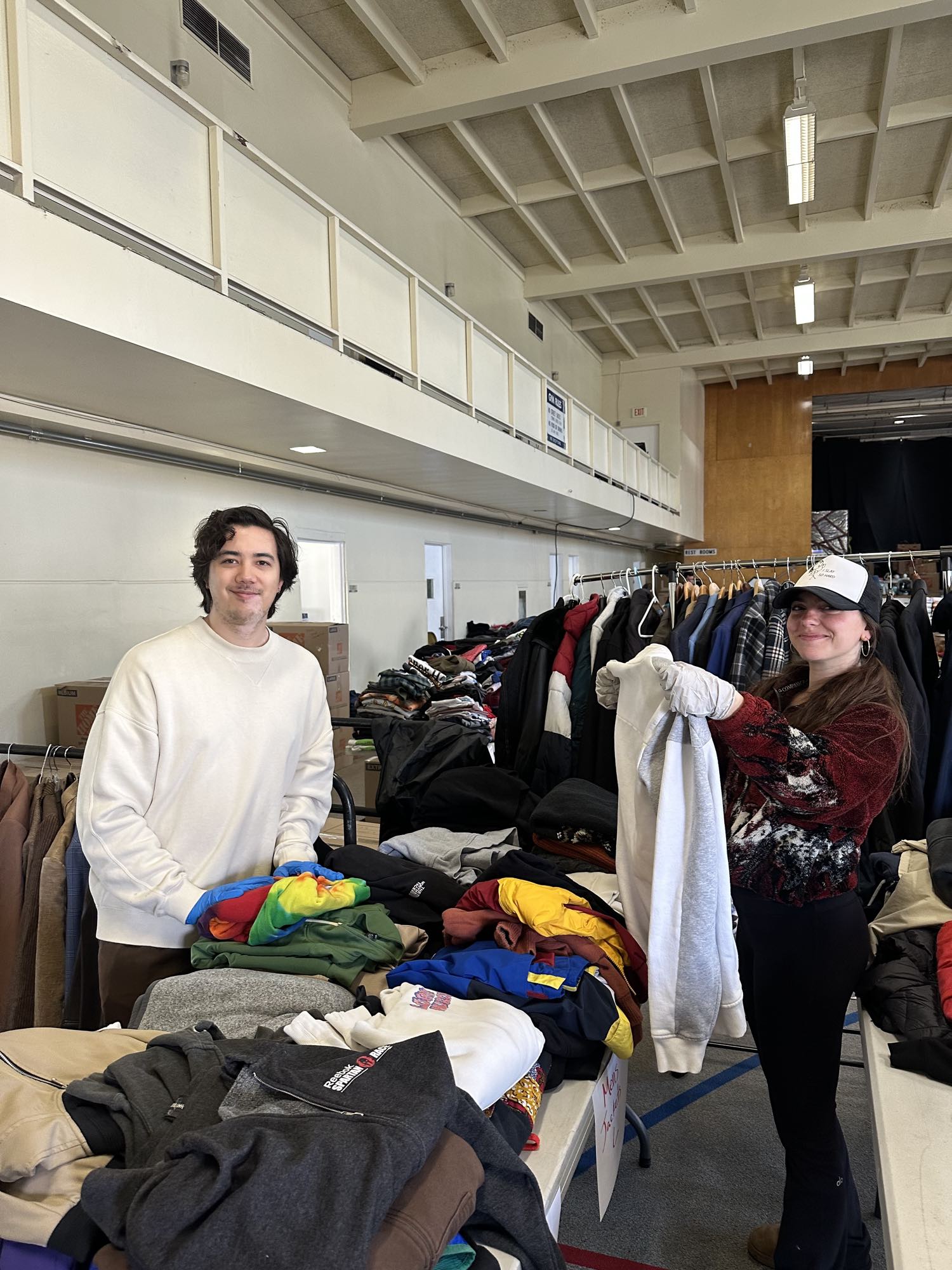 Two volunteers standing at clothing tables in a gymnasium, one behind stacks of folded sweatshirts and the other holding up a grey hooded sweatshirt while wearing work gloves, with a rail of hanging jackets between them
